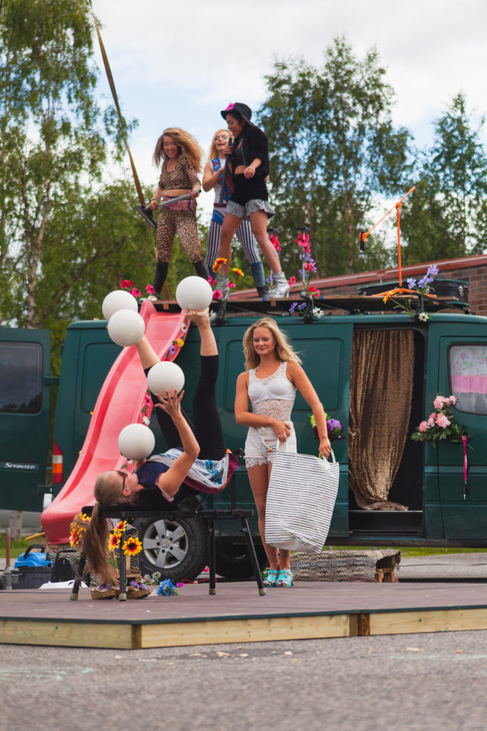 Viisi naista esittää sirkustaidetta vihreän pakettiauton ympärillä. Naisilla on värikkäitä esiintymisasuja. Pakettiauton katolta tulee alas liukumäki. Yksi naisista on selällään ja jongleeraa käsin ja jaloin viidellä pallolla yhtä aikaa.