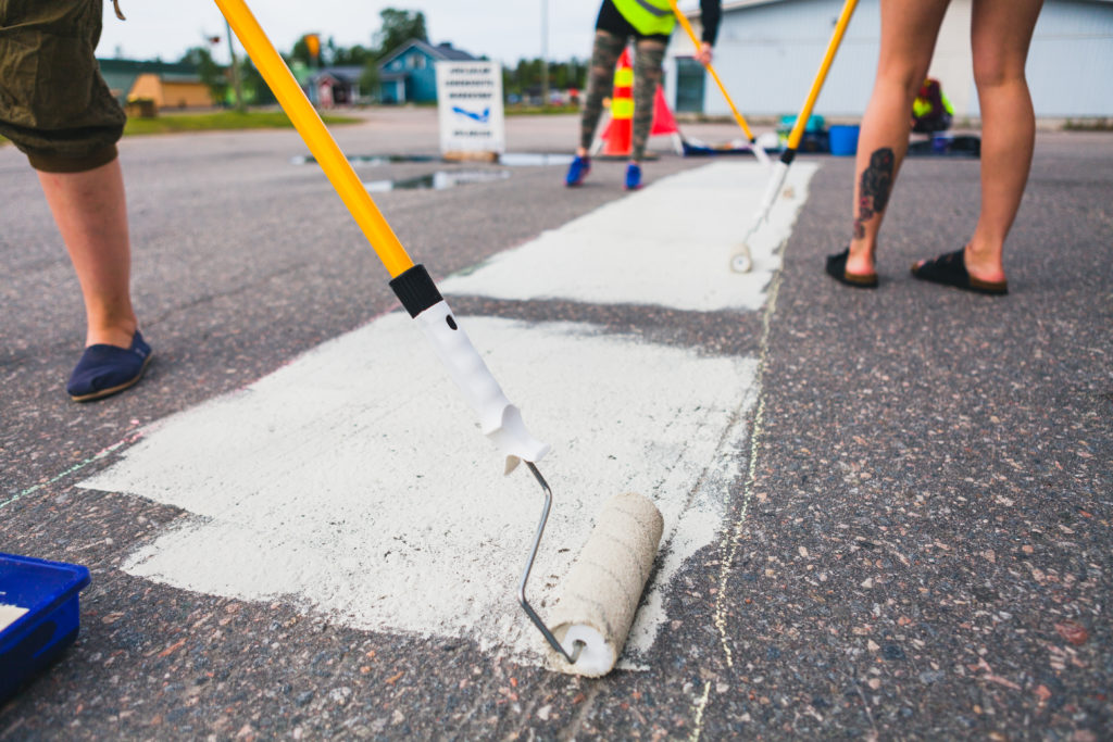 Kolme ihmistä maalaa asfalttiin mattoa valkoisella värillä varsitelalla katutaide-kurssilla Kolarissa.